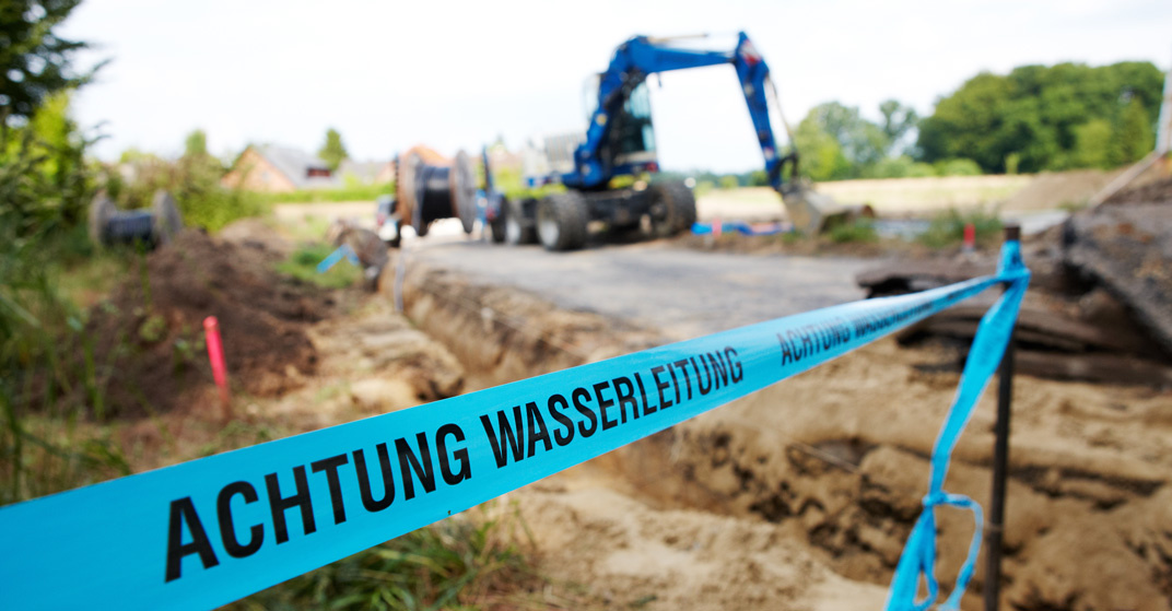 Blaues Absperrband mit der Aufschrift "Achtung Wasserleitung" vor einer Baustelle, auf der ein Bagger gräbt.