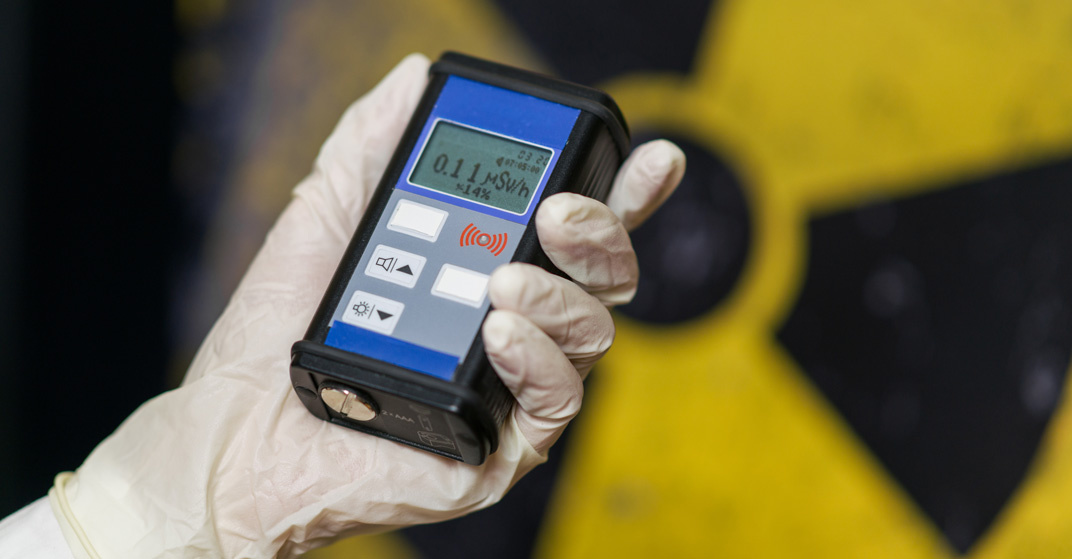 Hand mit Schutzhandschuh hält blauen Geigerzähler mit Messwert 0.11 vor gelbes Atomkraft-Symbol