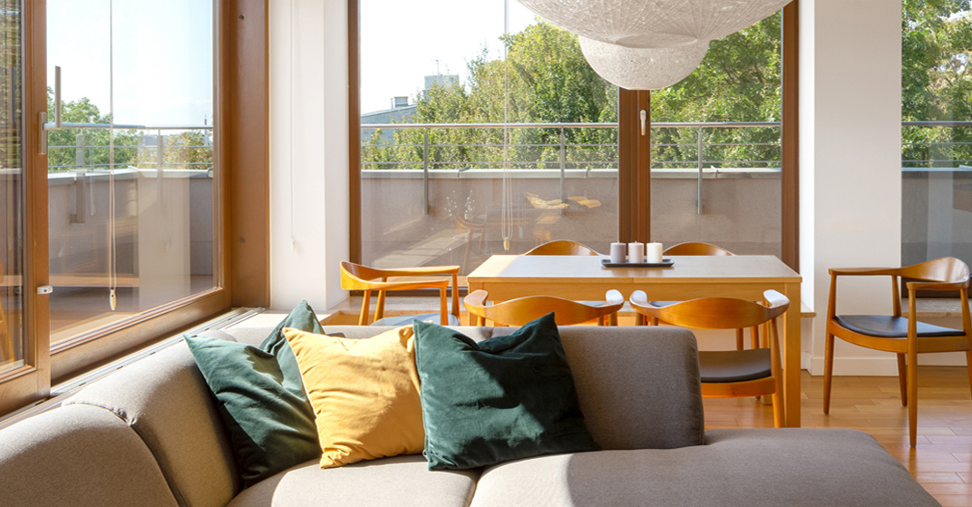 Wohn- Esszimmer mit großer Fensterfront mit bodenhohen Holz-Alu-Fenstern, graue Couch mit gelben und grünen Zierkissen, angenehmes Ambiente