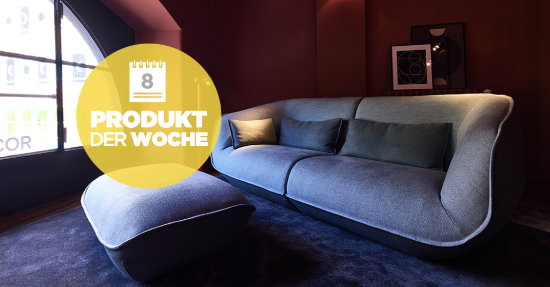 Sofa Nook von COR in Grau auf dunkelblauem Teppichboden vor weinrot gestrichener Wohnzimmerwand, Hocker von Nook steht davor